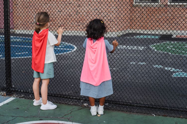 Two children wearing capes look through a playground fence, embracing imagination.
